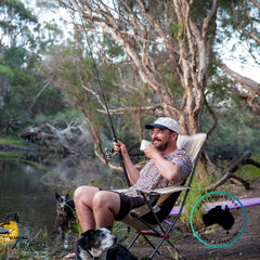 Man fishing by a river while sitting comfortably in a beige Dhubite Outdoors camp chair, holding a cup and smiling with his dogs nearby.