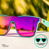 Knockaround Afters Miami Sunglasses with pink lenses and teal frames on a blurred natural background