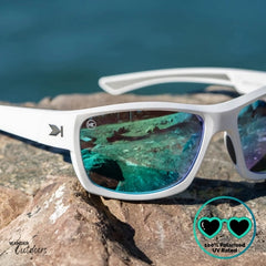 Knockaround High Stakes Aura Sunglasses with white frames and blue-tinted lenses on a rock with water in the background.