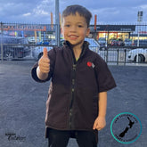 A young boy smiling and giving a thumbs up while wearing the Stoney Creek Kids Rammer Jacket in dark brown. The jacket features a high collar, front zip, and short sleeves. Wander Outdoors logo and New Zealand Owned & Operated badge visible.