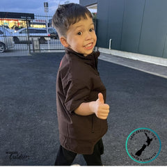 Side view of a boy wearing the Stoney Creek Kids Rammer Jacket in dark brown, smiling and giving a thumbs up. Shows jacket’s short sleeves, high collar, and side fit. Wander Outdoors and New Zealand Owned & Operated branding displayed.