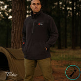 A middle-aged man wearing a black Stoney Creek Quarter Zip Top stands near a campsite at dawn. A swag and campfire are visible in the background, with soft morning light highlighting the rugged outdoor setting.