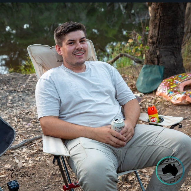 Camper relaxing in a beige Dhubite Outdoors compact chair with a drink in hand, showing armrests, dual cup holders, and comfortable seating angle.