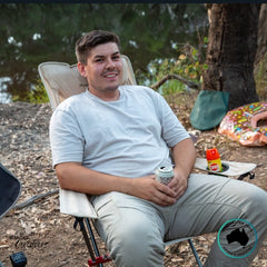 Camper relaxing in a beige Dhubite Outdoors compact chair with a drink in hand, showing armrests, dual cup holders, and comfortable seating angle.