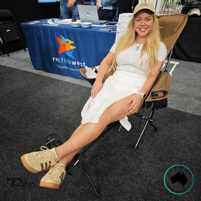 Woman reclining in a tan Dhubite Outdoors Compact Camp Chair with matching adjustable foot rest attached, demonstrating full-length comfort and relaxed seating posture.