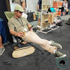 Man sitting in an olive green Dhubite Outdoors Camp Chair with attached foot rest, enjoying a drink and scrolling his phone at an indoor event.