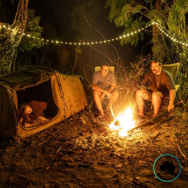 Campfire scene with Dhubite retractable string lights illuminating the campsite, showing warm even light strung between trees above swags and camp chairs.