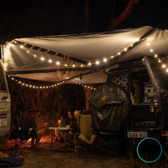 Dhubite retractable string lights set up under a 4WD awning, providing soft warm light for campers sitting around a table at night.