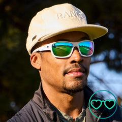 Man wearing a beige cap and Knockaorund High Stakes Sunglasses in Aura with a blurred natural background playing golf.
