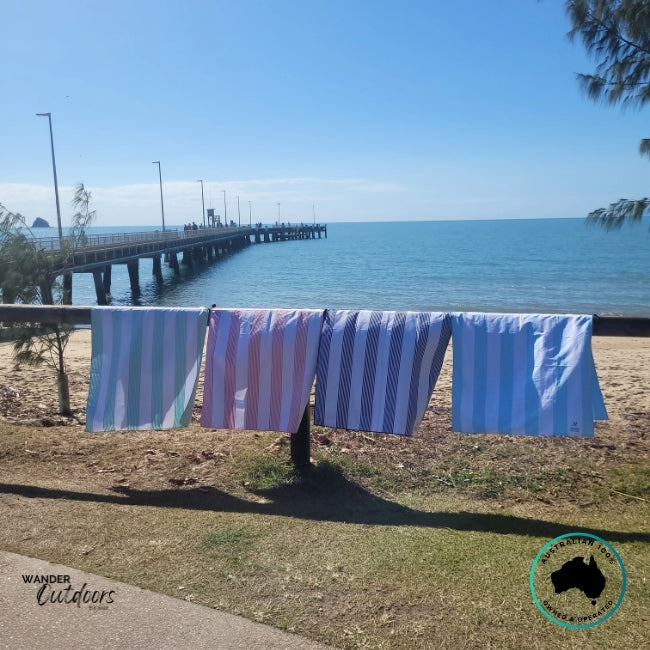 Newlyfe Signature Beach Towels in Calvi Sky Blue, Sanur Pink, Milos Mint and Amalfi Navy hanging along a beachside fence overlooking the ocean and jetty.
