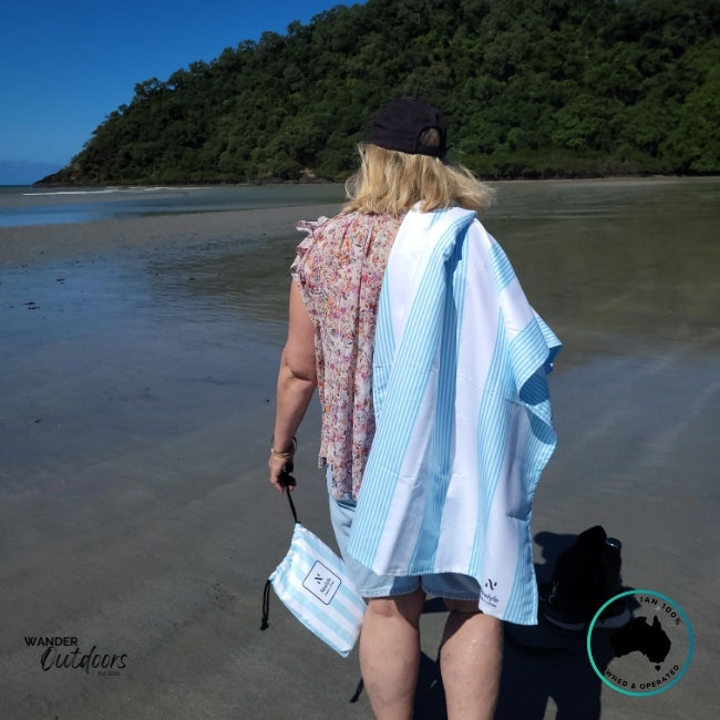 Person standing on a tropical beach holding the Newlyfe Signature Calvi Sky Blue Towel and matching pouch over their shoulder.