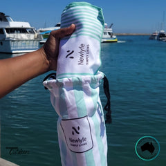Hand holding the Newlyfe Signature Milos Mint Sand-Free Beach Towel rolled into its matching striped pouch beside the marina.