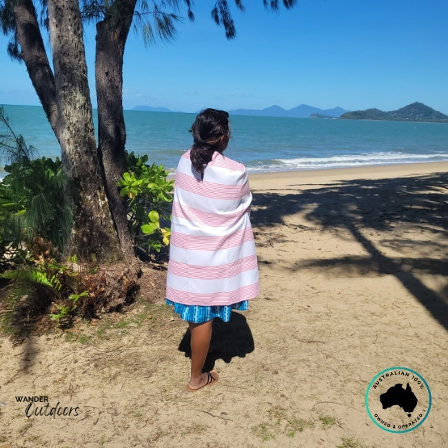 Person wrapped in Newlyfe Signature Sanur Pink Towel while standing under trees near the shoreline.