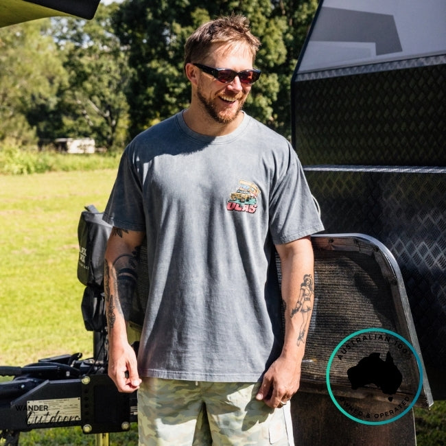 Man wearing a gray t-shirt with a logo, standing next to a caravan in an outdoor setting.