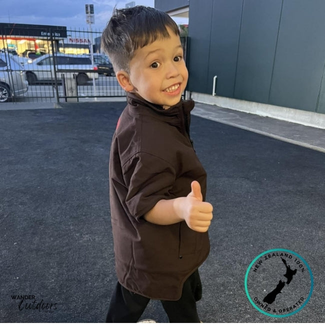 Side view of a boy wearing the Stoney Creek Kids Rammer Jacket in dark brown, smiling and giving a thumbs up. Shows jacket’s short sleeves, high collar, and side fit. Wander Outdoors and New Zealand Owned & Operated branding displayed.