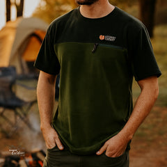 Person wearing a green and black fleece t-shirt with a logo, standing outdoors.