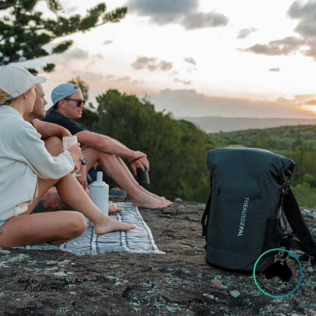 three people sitting on a rock with a scenic view, an Aussie Pal backpack  in the foreground.