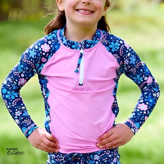 A young girl wearing a long-sleeve rashie with a bright pink front panel and blue floral-patterned sleeves, standing outdoors with her hands on her hips and smiling. The rashie has a half-zip at the neckline.