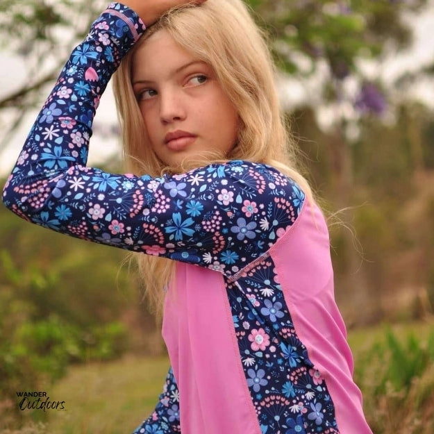 A girl with long blonde hair wearing the same rashie, posing with one arm raised. The side view showcases the floral-patterned sleeves and side panels, with the pink front and back sections. The background is a natural, outdoor setting.