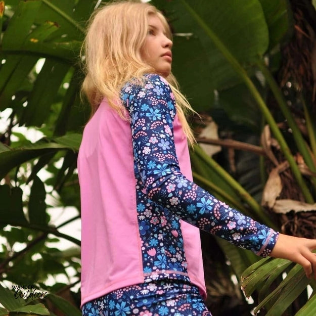 The same girl is captured from the side, slightly looking away. The image highlights the full length of the rashie’s sleeves and body, showing the contrast between the vibrant floral pattern and the solid pink panels. The background is lush with greenery.