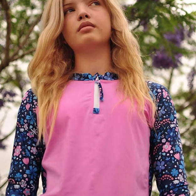 A older girl wearing a long-sleeve rashie with a bright pink front panel and blue floral-patterned sleeves, standing outdoors with her hands on her hips and smiling. The rashie has a half-zip at the neckline.