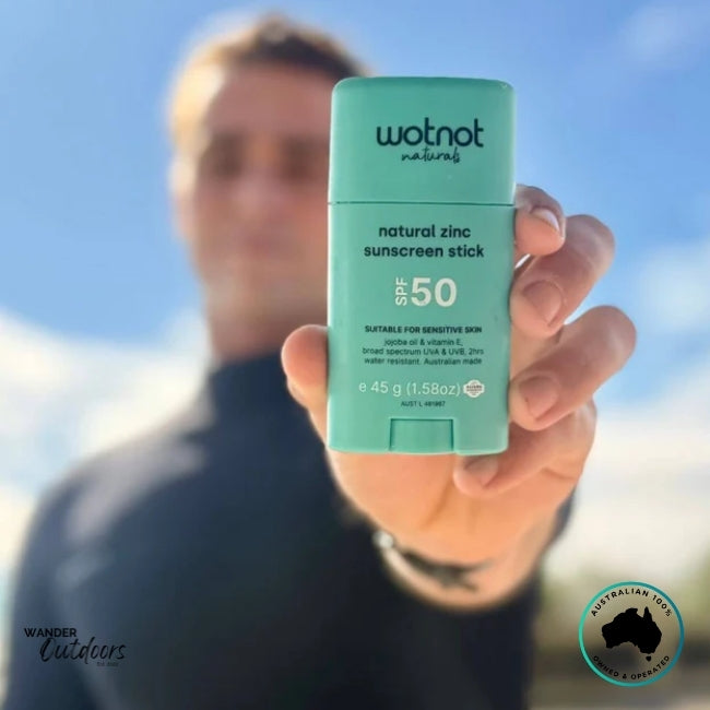 Surfer holding Wotnot Naturals SPF 50 Zinc Sunscreen Stick toward camera with blue sky and surfboard in background.
