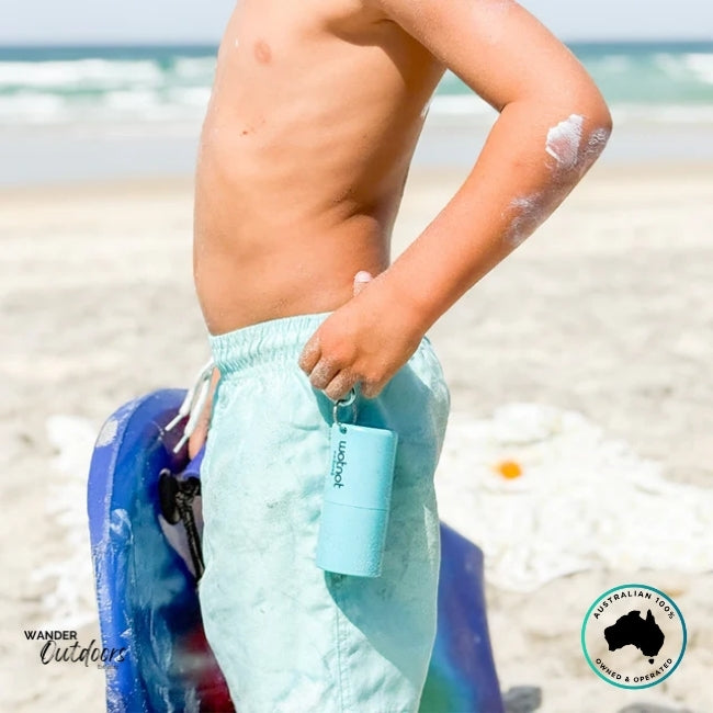Child standing on the beach with Wotnot Refillable Sunscreen Roll-On clipped to swim shorts while applying sunscreen